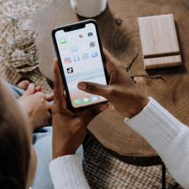 A person is using an iPhone with various app icons visible on the screen, including TikTok, Instagram, and Facebook, while seated at a wooden table—things to think about if you're a marketer on TikTok.