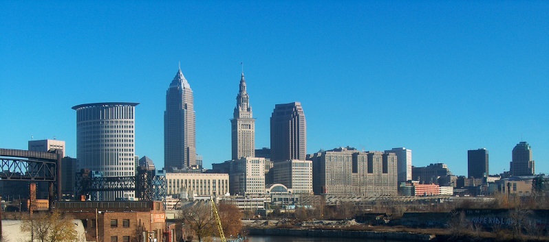 Skyline of Downtown Cleveland with predominantly modern high-rise buildings and some historic structures, viewed from an elevated perspective under a clear blue sky.