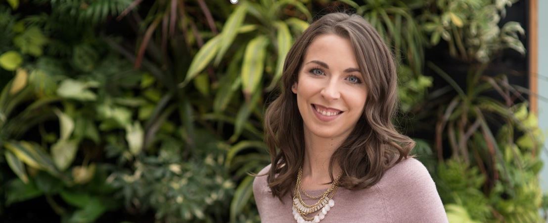 Hannah Wineland, a woman with shoulder-length brown hair, wearing a light purple sweater and a layered necklace, smiles in front of a background of various green plants.