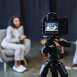 A camera on a tripod is focused on a person seated in a chair, with the scene being filmed visible on the camera's screen, capturing essential media interview moments.