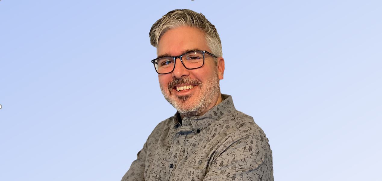 Tom Tennant, a Senior Content Strategist with gray hair and glasses, smiles while wearing a patterned shirt against a light blue background.