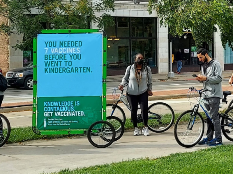 A sign on a bicycle trailer reads, "You needed 7 vaccines before you went to kindergarten. Knowledge is contagious. Get vaccinated." People with bicycles are nearby, promoting the CSU message: Facts Matter.