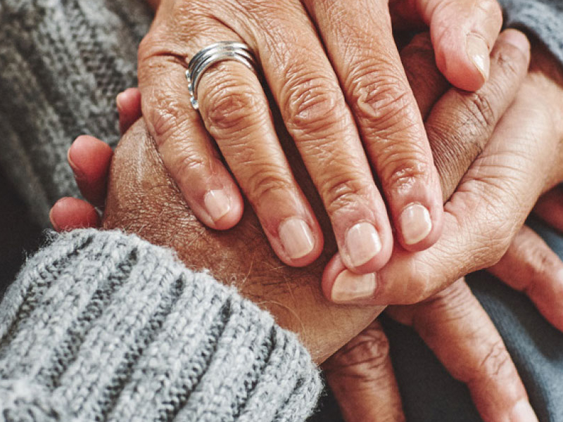 Two pairs of hands in a close embrace, with one pair wearing a gray knitted sweater and the other adorned with a silver ring, exuding an air of care and tenderness.