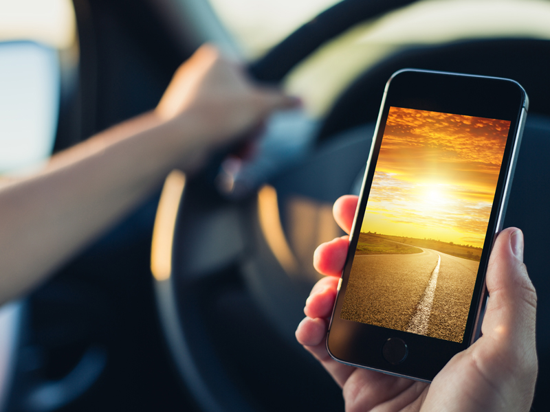 A person holds a smartphone displaying a sunset over a road, with their other hand on a steering wheel, suggesting they are in a car—perhaps considering the peace of mind that comes with Erie Insurance.