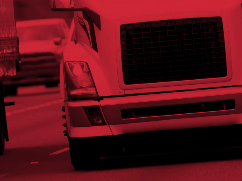 Close-up of the front of a large truck on a road, with a red filter applied to the image. The truck, equipped with Federal Mogul parts, dominates the scene while other vehicles are visible in the background.