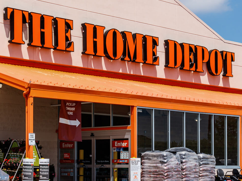 Exterior of The Home Depot store with signage, orange awning, and entrance doors visible. Stacks of products are in front of the Depot.