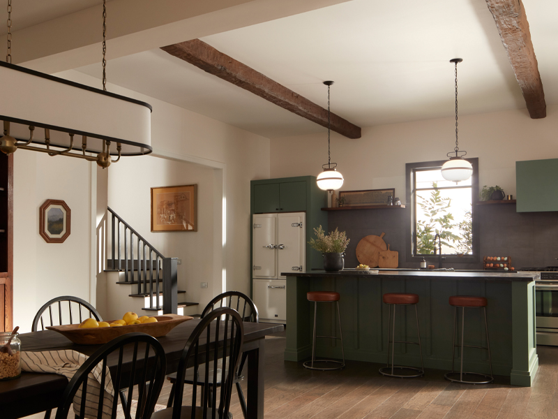 A modern kitchen and dining area with wooden beams, black chairs, a green island with bar stools, pendant lights by Kichler, dark gray backsplash, and various kitchen accessories. There's a wooden staircase in view.