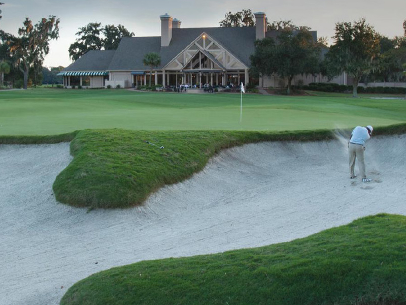 A golfer at The Landings attempts to hit a ball out of a sand bunker on a pristine golf course, with the elegant clubhouse and towering trees visible in the background.