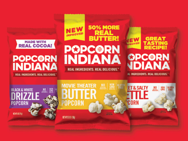 Three bags of Popcorn Indiana are displayed: Black & White Drizzle, Movie Theater Butter, and Sweet & Salty Kettle. Each Indiana popcorn bag highlights specific features and ingredients against a vibrant red background.