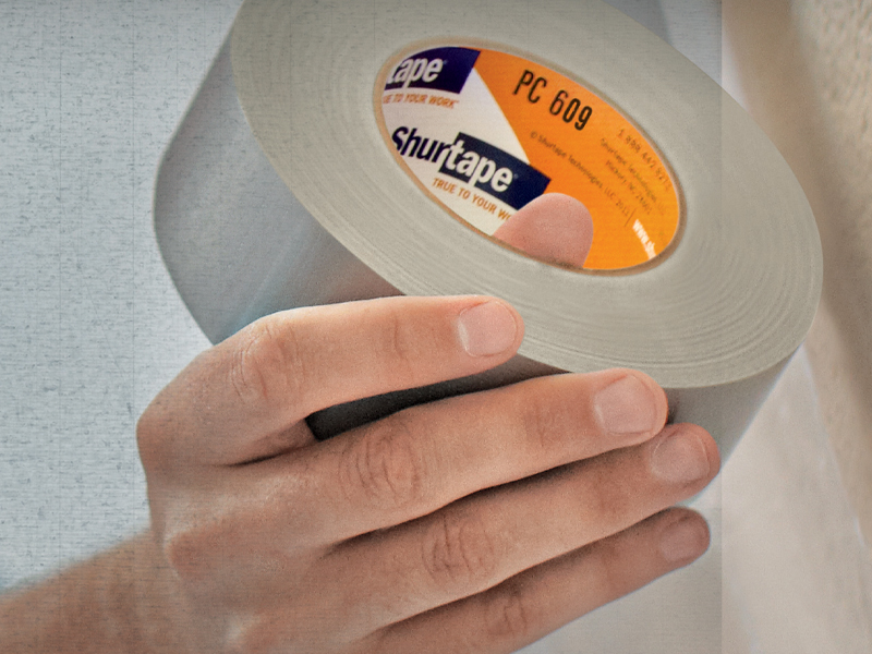 A close-up of a hand holding a roll of Shurtape PC 609 cloth tape, an essential in industrial solutions, with gray adhesive tape visible.