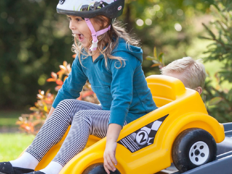 A young child wearing a helmet sits in a yellow toy car, looking excited, just like the ones featured on the Step2 website. Another child is partially visible behind them as they enjoy the outdoors with lush greenery in the background.