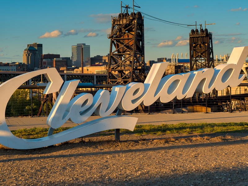 A large sign displaying "Cleveland" in script font is placed in front of an industrial backdrop and the Downtown city skyline during sunset.