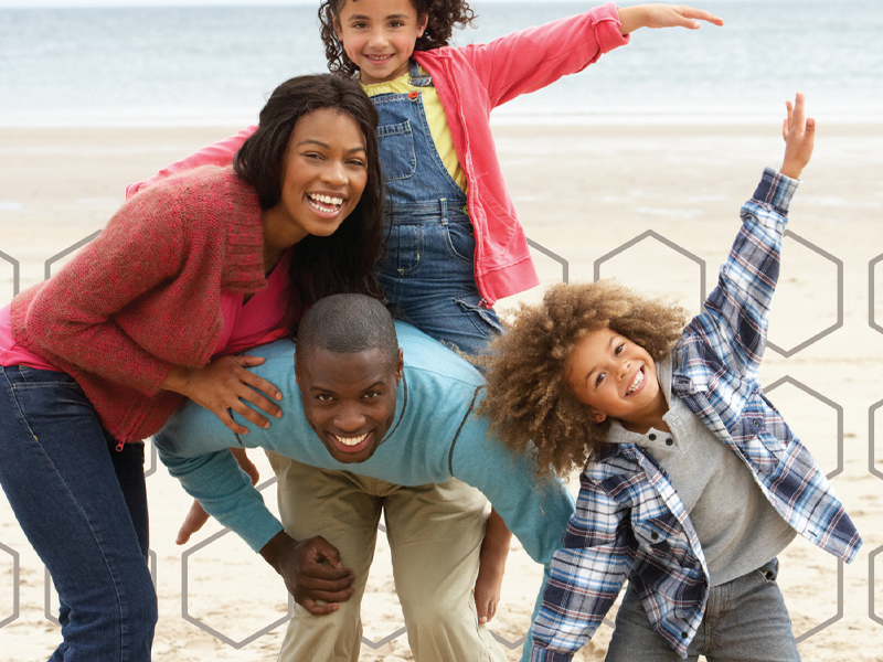 A smiling family of four poses playfully on a beach, with the mother and daughter standing behind the father, who is crouched down, and the son standing next to him, arms outstretched—much like their last vacation funded by Huntington Bank.