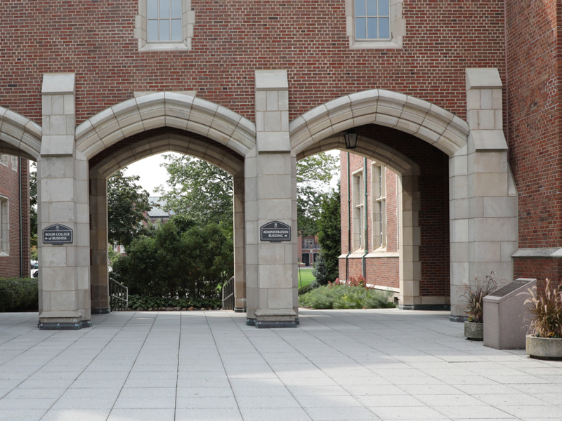 Two stone archways with signage indicating the Rose Garden and Memorial Garden, part of a brick building with sidewalks and greenery in the background at John Carroll University.