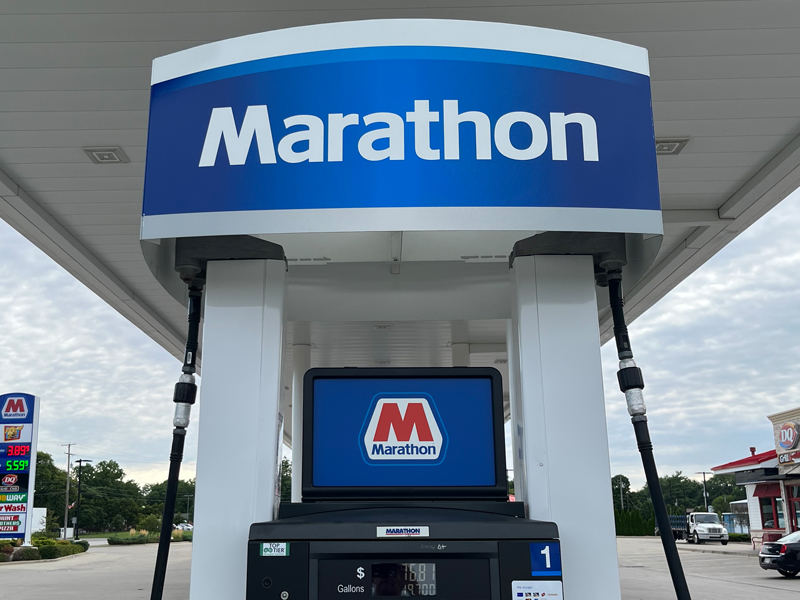 A gas pump at a Marathon gas station with the Marathon logo prominently displayed on the canopy and pump, much like a runner showcasing pride in a long-distance race. Gas prices are shown on a sign in the background.