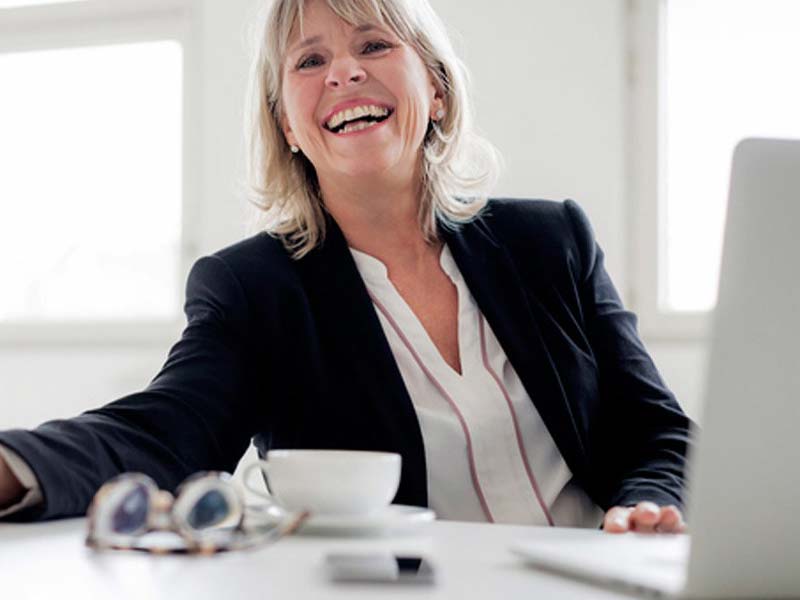 A smiling woman with blonde hair sits at a desk with a laptop displaying the NAMS logo, a cup, and glasses in the foreground.