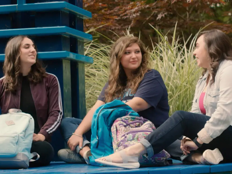 Three women sit outdoors on blue steps at Northwood University, engaging in conversation. They have backpacks with them and are surrounded by greenery, exemplifying the quintessential university experience.