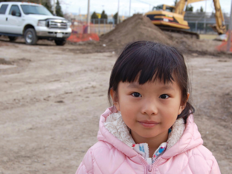 A young girl wearing a pink jacket stands on a construction site with a white truck and an excavator in the background