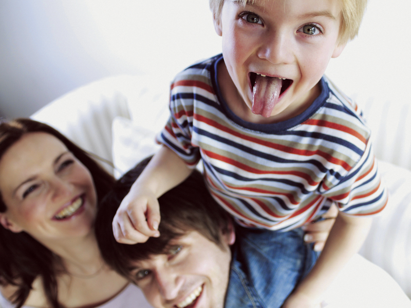 Child in a striped shirt playfully sticks out tongue while sitting on adults' shoulders; all smiling and appearing joyful. Nearby, a bottle of Purell hand sanitizer ensures their hygiene remains in check as they enjoy their fun-filled day.