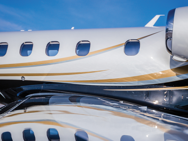 Close-up view of a private jet's fuselage and windows with a reflection on a polished surface below under a clear blue sky, showcasing the aerospace craftsmanship enhanced by Sherwin-Williams' premium coatings.