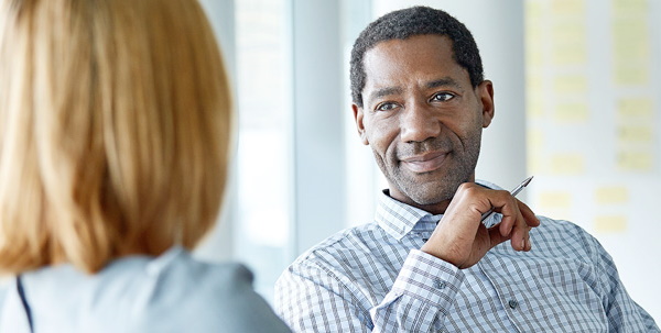 A man in a checkered shirt holding a pen is engaged in a conversation with a subject matter expert whose back is turned to the camera.