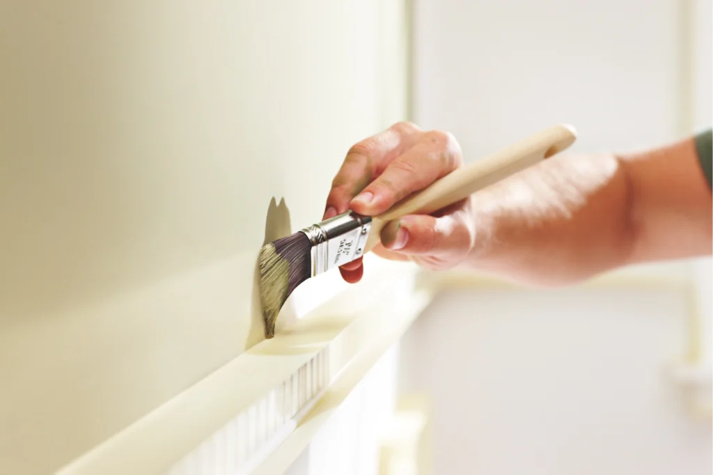 A person uses a paintbrush to apply light-colored paint to an interior wall above white trim.