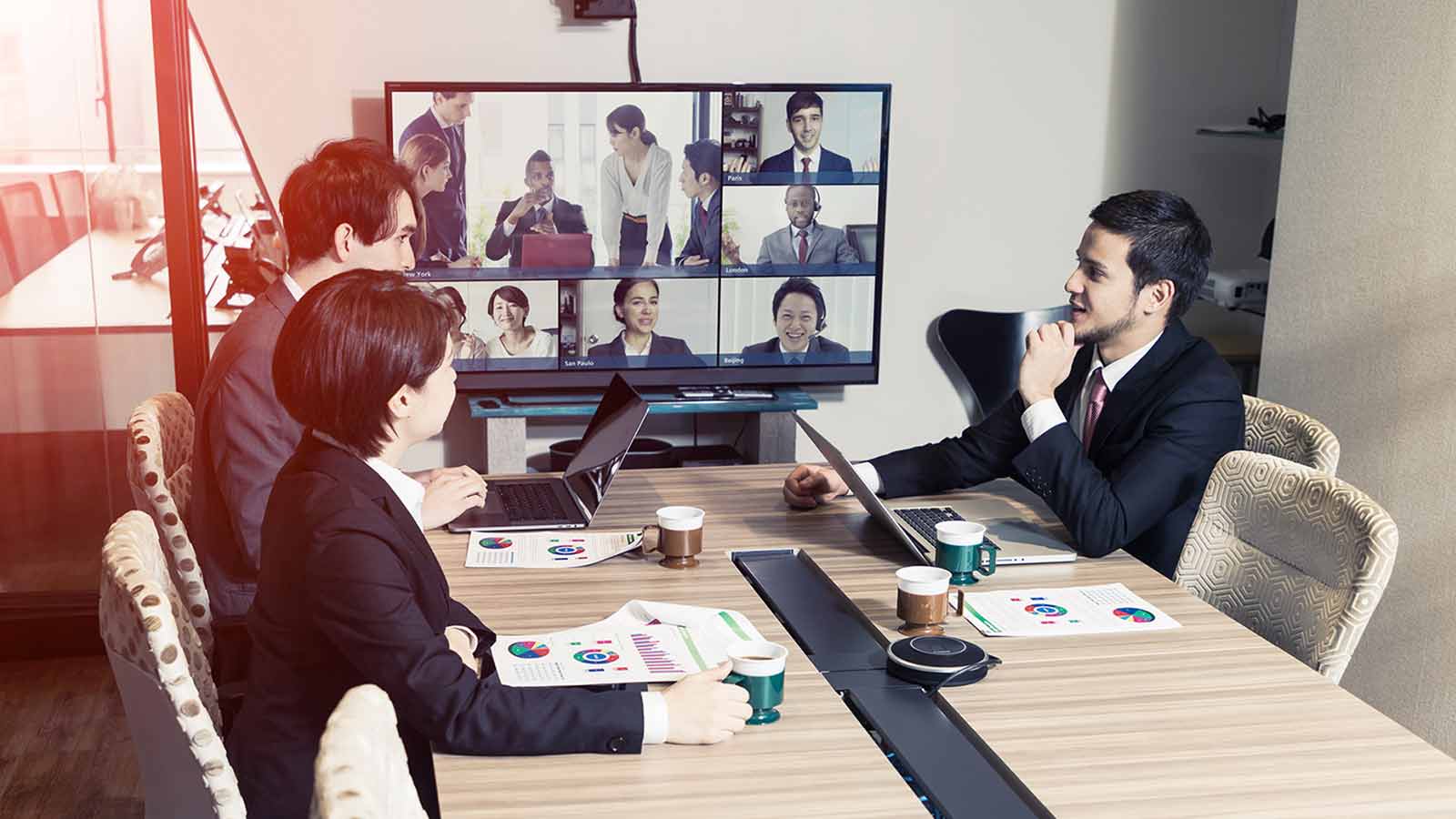 Three people in a conference room participate in a virtual meeting with several others shown on a screen, exemplifying the future of events. They are dressed in business attire and have laptops and documents in front of them.