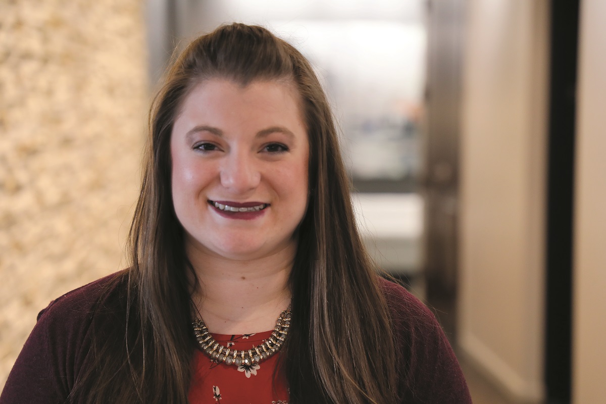 Annie Morino, a member of the Falls & Co. research team, smiles in a hallway with long brown hair, wearing a maroon cardigan and a statement necklace against a blurred background.