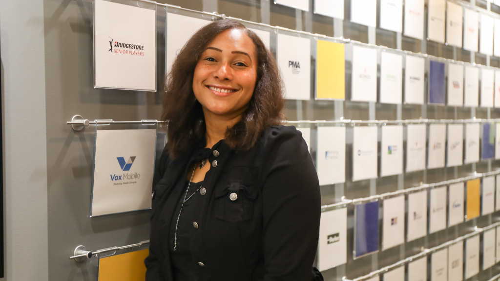 A person wearing a black jacket stands smiling in front of a wall featuring various company logos displayed in clear panels; this is Chante Jones, Associate Vice President of Communications.