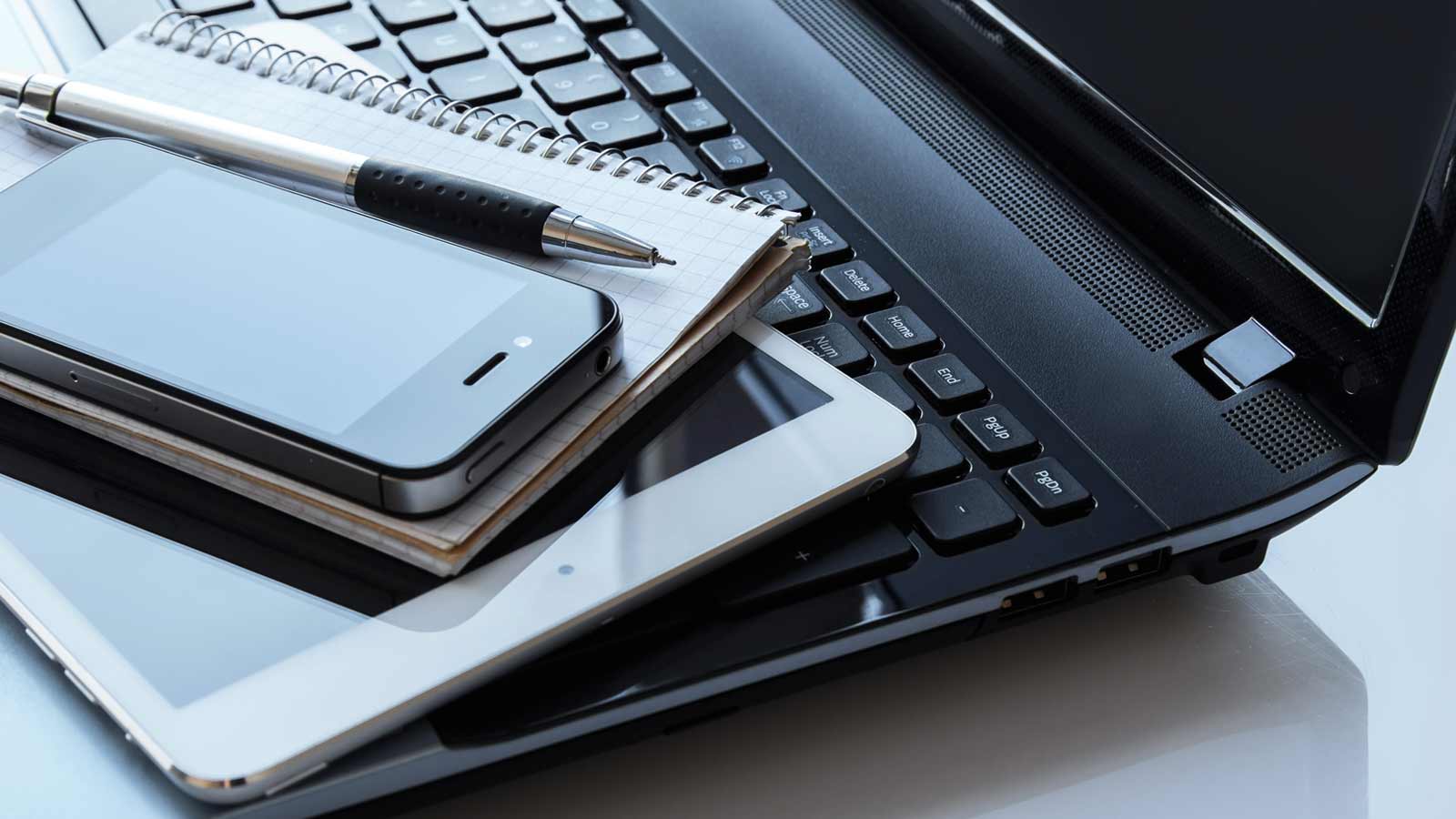 A laptop with a black keyboard has a smartphone, tablet, notebook, and pen placed on it, serving as a consistent reminder of the tools essential for staying connected and organized.