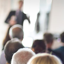 Audience members seen from behind are engaged in a session of corporate storytelling as the speaker artfully commands attention at the front of the room.