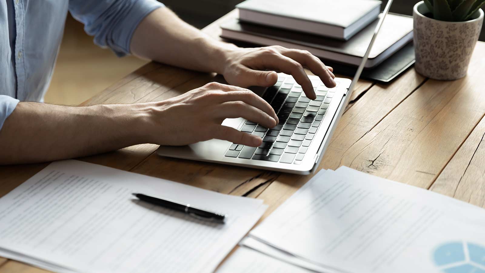 Person typing on a laptop at a wooden desk with documents and a pen, skillfully crafting blog posts to enhance SEO.