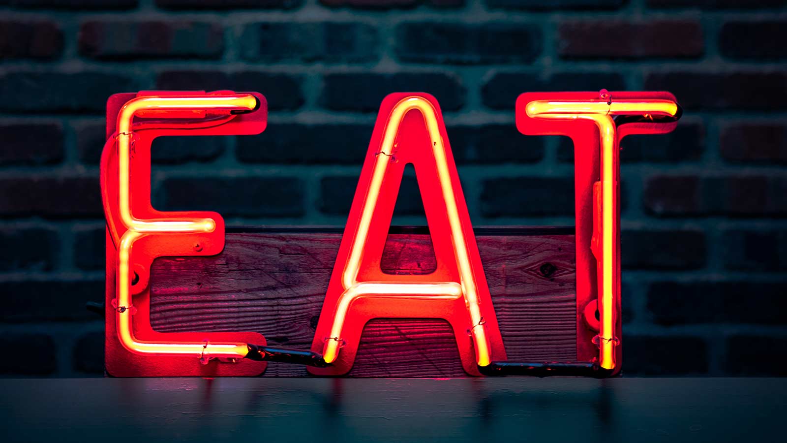 Neon sign reading "EAT" mounted on a wooden board against a brick wall background, evoking the principles of E.A.T. SEO for trusted website content.