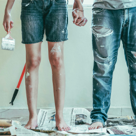 Two barefoot individuals in paint-splattered clothing, holding hands, stand on a newspaper-covered floor with a paintbrush amidst painting tools and supplies, dreaming of homeownership despite high home prices.