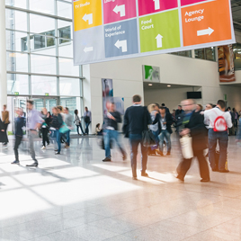 People walking through a busy indoor area with a high ceiling and large windows. Overhead signs indicate directions to various sections, including TV Studio, World of Experience, Food Court, and Agency Lounge. The layout suggests an effective trade show marketing strategy like those seen at the NAHB International Builders' Show.