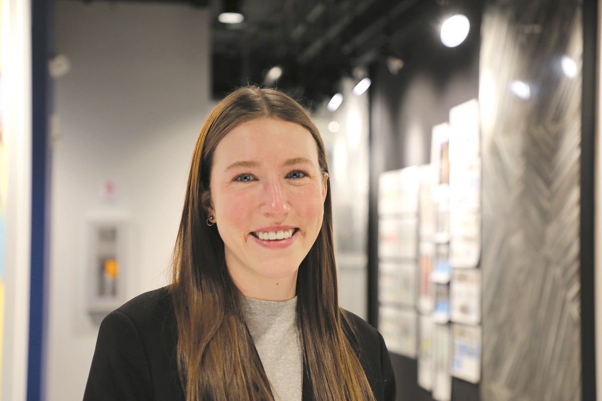 Kiersten Becht, a member of the Communications Team at Falls & Co., smiles at the camera in a well-lit indoor setting. She has long brown hair and is wearing a blazer over a gray shirt, with blurred background elements