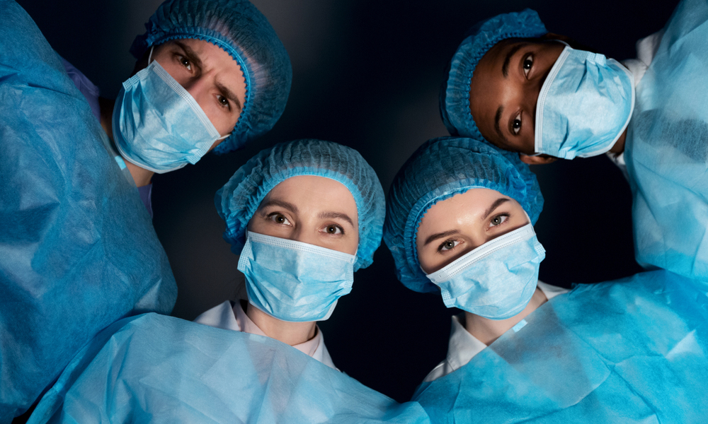 Four medical professionals wearing blue surgical gowns, hair covers, and face masks, looking directly down at the camera, suggesting a perspective of someone lying on an operating table or examination bed—an image meticulously refined through crowdsourced editing.