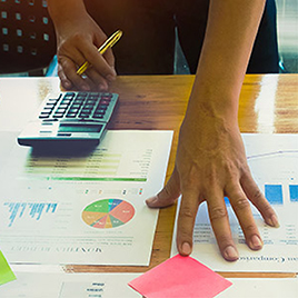 A person holding a yellow pen uses a calculator while reviewing financial documents and charts, along with colorful sticky notes, to measure PR program performance.