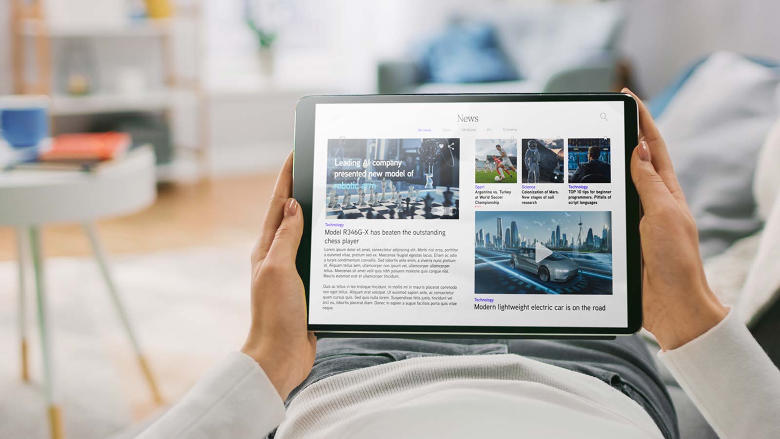 Person holding a tablet displaying a news website with articles on various topics such as economy, technology, and sports. The media coverage is comprehensive. A living room in the background.