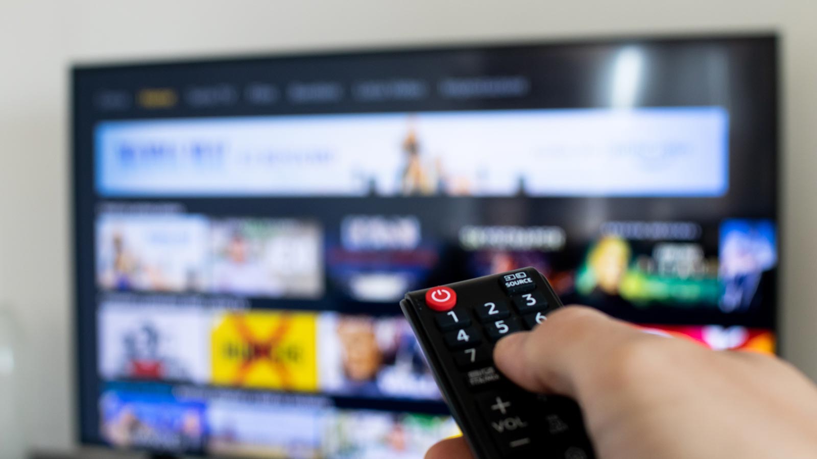 A hand holds a remote control pointed at a television displaying a streaming service interface with various show and movie options, illustrating the ongoing viewership shift from traditional cable television.