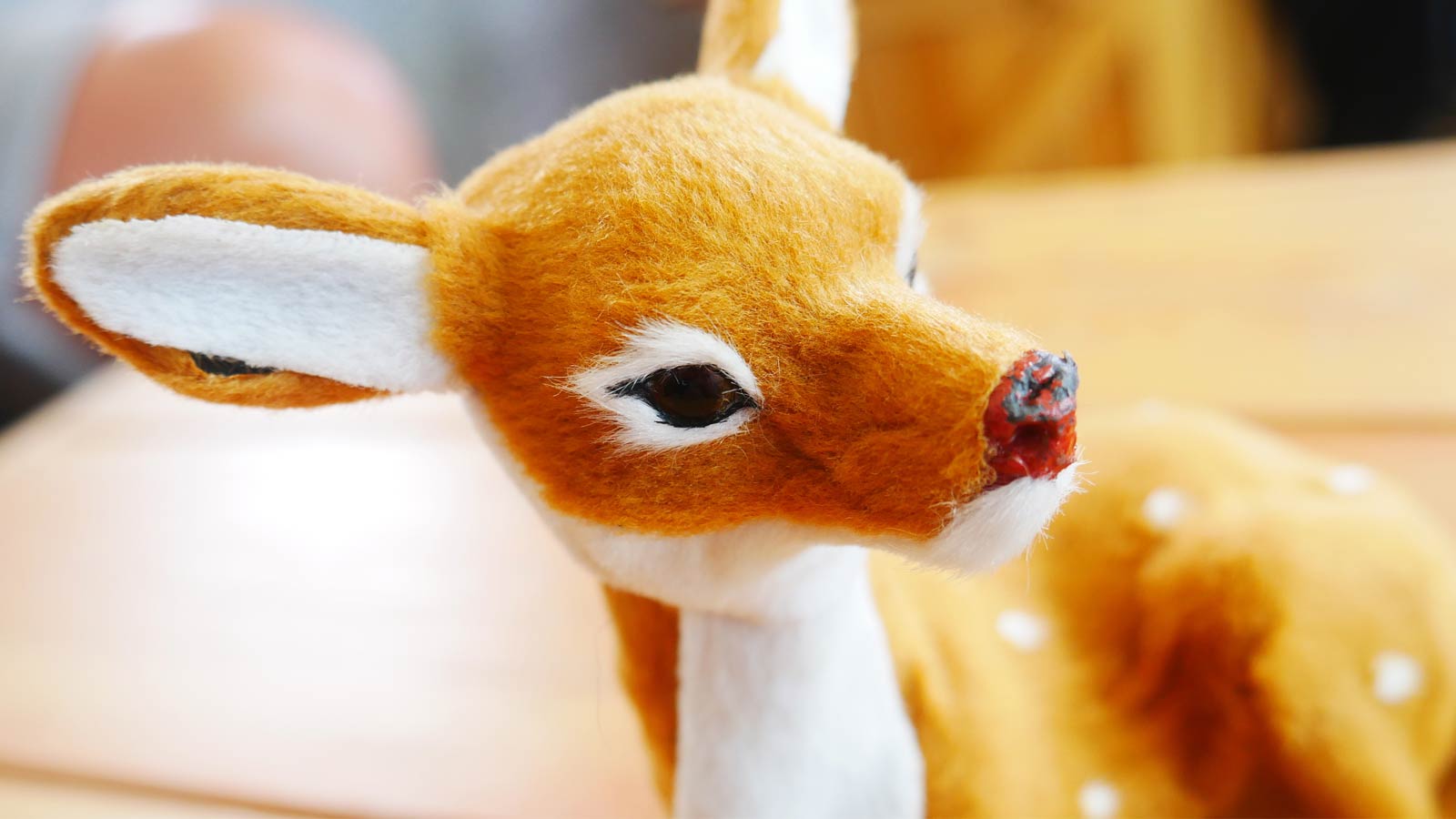 A close-up image of a small, plush toy deer with brown and white fur, focusing on the deer's face and head, positioned on a wooden surface adorned with twinkling Christmas lights.