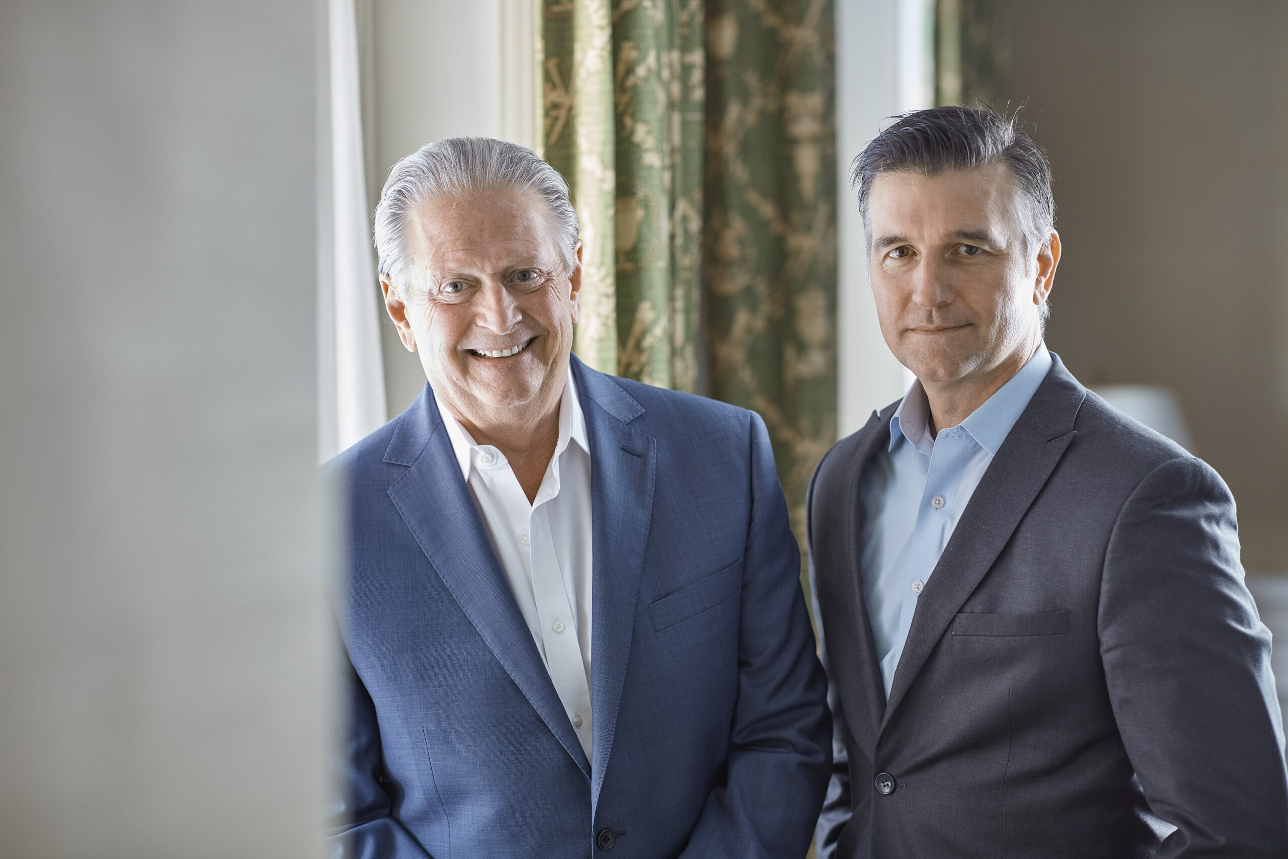 Two men in business attire stand side by side indoors, both wearing blue suits and white shirts. The older man on the left, identified as Tom Bernot, President, is smiling while the younger man on the right has a neutral expression.