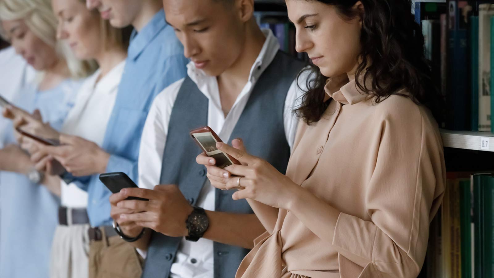 A group of people stands in a line near bookshelves, each focused on their smartphones, undoubtedly checking out the latest social media strategy.