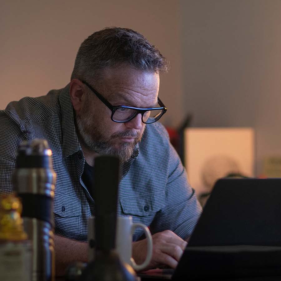 A man with glasses and a beard is intensely focused on his laptop screen, sitting at a table with a coffee mug and thermos, perhaps analyzing Super Bowl commercials.