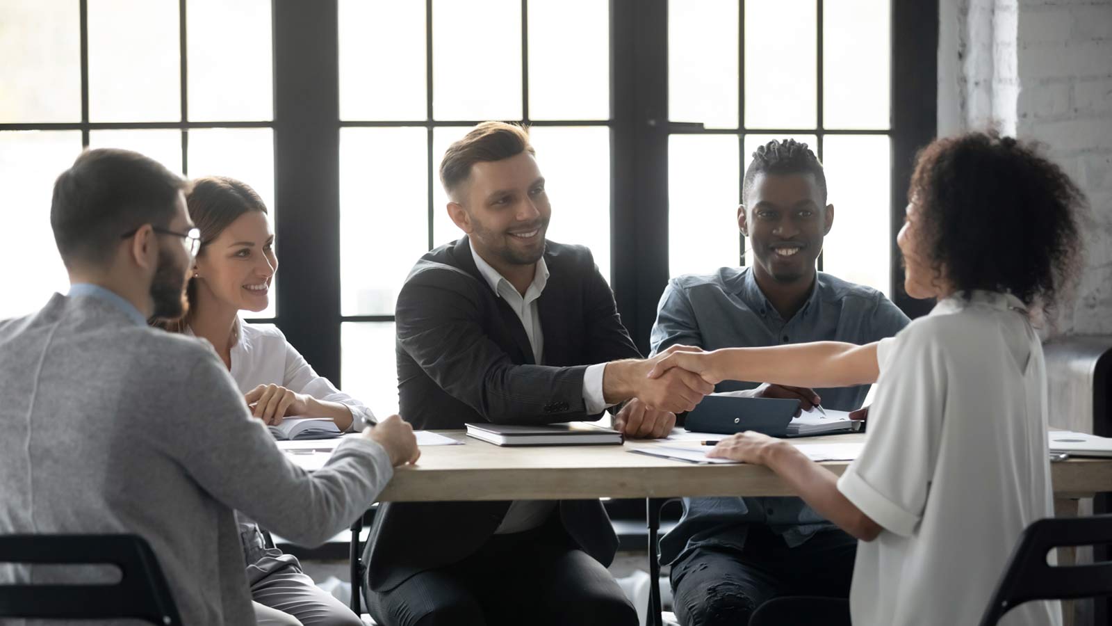 A group of five people sit around a table in an office, with two people in the center shaking hands, solidifying a strong client-agency relationship.