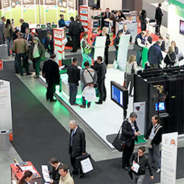 A busy exhibition hall with multiple booths, attendees walking around, and people engaging with exhibitors. Various displays and informational stands are prominently featured, capturing the essence of major trade shows.