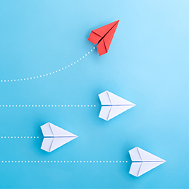 Four paper planes in flight, with one red plane veering upwards while three white planes fly straight below it, all against a blue background—a vivid metaphor for a successful Public Relations Strategy.