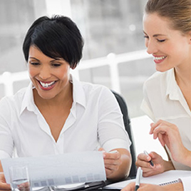 Two women in white shirts sitting at a table. One woman is holding a paper and smiling, while the other woman is looking at the paper and also smiling, as if discussing how to work with an agency.