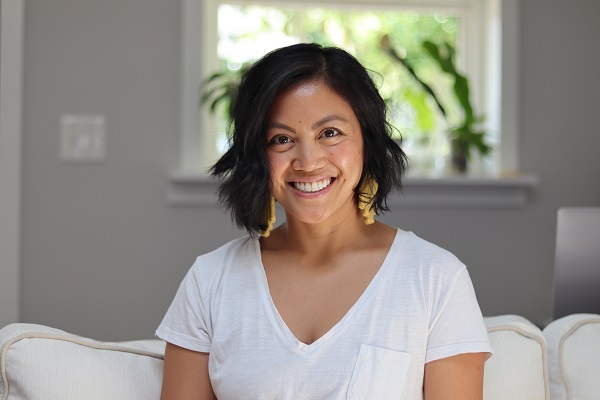 A person with short, dark hair and a white shirt smiles while sitting on a light-colored sofa. Justine DeJesus, the Social Media Specialist at Falls & Co., is perfectly framed by a window with plants behind her.