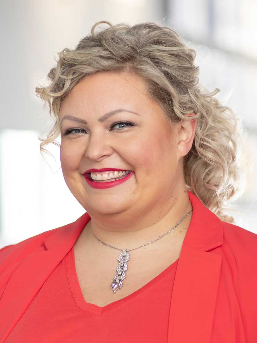 Kristin Pohlig, an Account Executive at Falls & Co., smiles warmly, her curly blonde hair framing her face as she dons a vibrant red blazer and a necklace with a pendant.